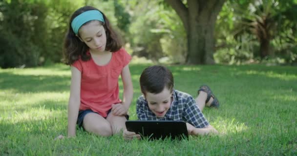 Um menino e uma menina deitados no gramado brincando com um computador tablet — Vídeo de Stock