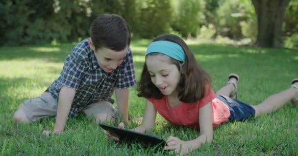 Een jongen en een meisje opleggen op het gazon met een tablet-computer spelen — Stockvideo