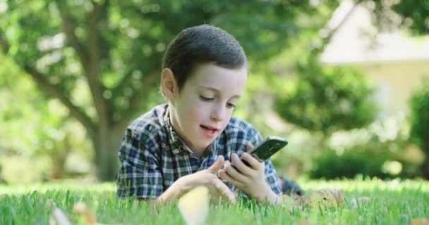 Мальчик играет с мобильным телефоном, лежащим на газоне — стоковое видео