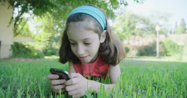 Uma menina brincando com um telefone celular deitado na grama — Vídeo de Stock