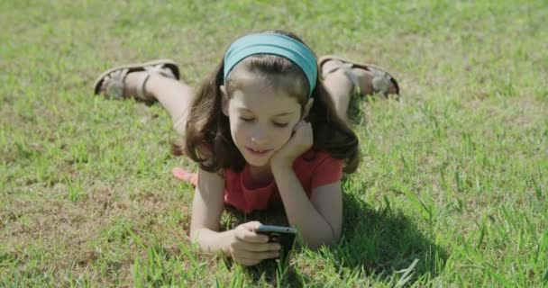 Una chica jugando con un teléfono móvil tendido en la hierba — Vídeos de Stock