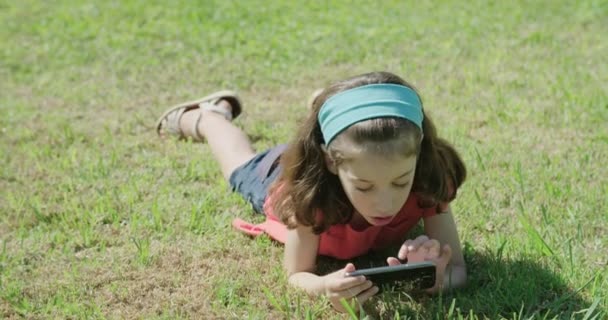 Девушка играет с мобильным телефоном, лежащим на траве — стоковое видео