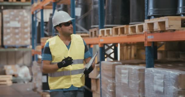 Tracking shot de un trabajador logístico que llevaba un casco inspeccionando artículos en un gran almacén — Vídeo de stock