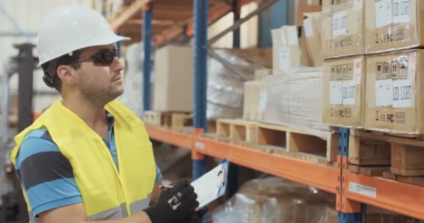 Tracking shot of a logistics worker inspecting items with a clipboard in a large warehouse — Stock Video