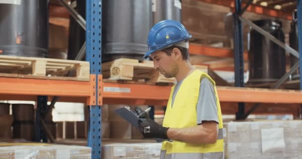 Tracking shot van een werknemer van de logistiek het dragen van een helm inspectie van artikelen in een grote magazijn — Stockvideo