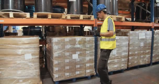 Tracking shot van een werknemer van de logistiek het dragen van een helm inspectie van artikelen in een grote magazijn — Stockvideo