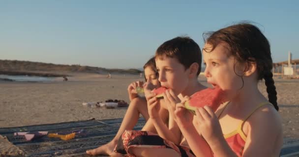 Tres niños comiendo sandías en la playa al atardecer — Vídeos de Stock