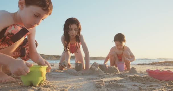 Tres niños construyendo castillos de arena en la playa al atardecer — Vídeos de Stock
