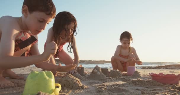 Tres niños construyendo castillos de arena en la playa al atardecer — Vídeos de Stock