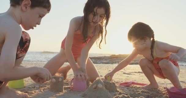 Трое детей строят песчаные замки на пляже во время заката — стоковое видео