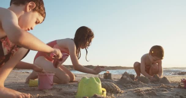 Tre bambini costruiscono castelli di sabbia in spiaggia durante il tramonto — Video Stock