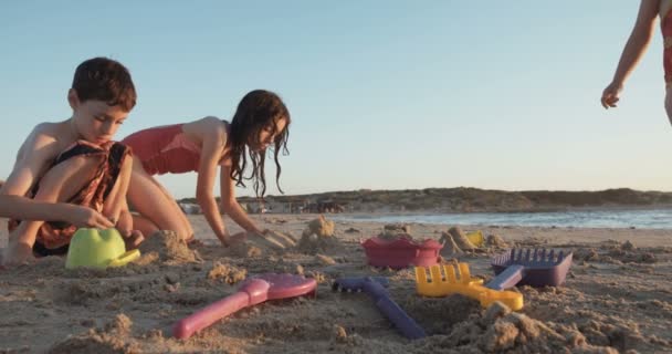 Tres niños construyendo castillos de arena en la playa al atardecer — Vídeos de Stock