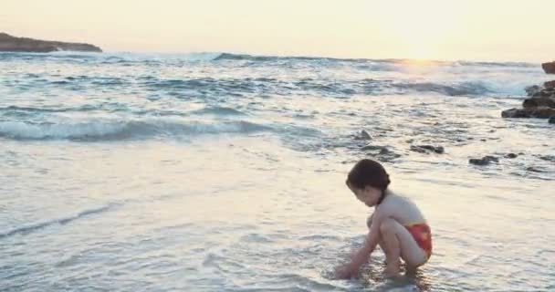 Kleines Mädchen sitzt am Strand und genießt die Wellen während des Sonnenuntergangs — Stockvideo