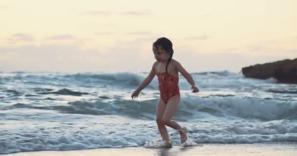 Mała dziewczynka siedzi na piasku na plaży, ciesząc się fale podczas zachodu słońca — Wideo stockowe