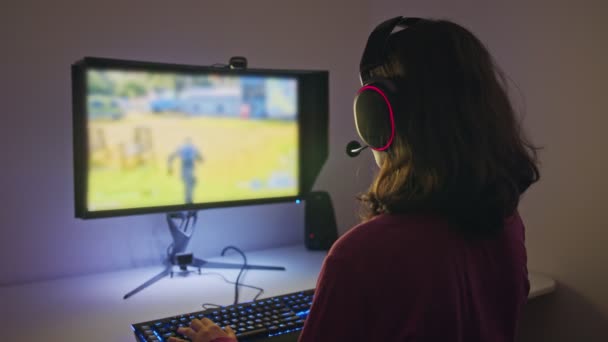 Ένα έφηβο κορίτσι που κάθεται μπροστά από έναν υπολογιστή, παίζοντας ένα παιχνίδι φορώντας ένα ακουστικό — Αρχείο Βίντεο