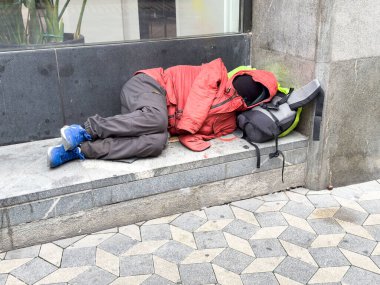 Ljubljana, Slovenia - October 10, 2025: Homeless individual resting on a bench in the city, wearing a red jacket, with a backpack nearby, showcasing urban challenges
