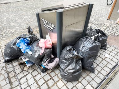 Ljubljana, Slovenia - October 10, 2025: Overflowing trash bin with black garbage bags on cobblestone street, showcasing urban waste issues and environmental impact