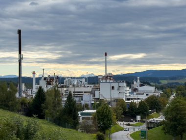 Althofen, Avusturya - 17 Eylül 2025: Endüstriyel fabrika bulutlu bir gökyüzüne karşı kurulmuş, üretim ve doğanın birlikteliğini ön plana çıkaran bacalar ve yeşillikler barındırıyor