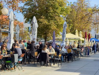 Graz, Avusturya - 11 Ekim 2025: Canlı sonbahar yapraklarıyla çevrili büyük şemsiyeler altında içeceklerin tadını çıkaran çeşitli insanlarla dolu açık hava kafesi