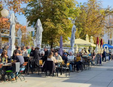 Graz, Avusturya - 11 Ekim 2025: Sonbahar ağaçlarıyla çevrili renkli şemsiyeler altında yemek yiyen ve sosyalleşen çeşitli insanlarla dolu bir açık hava kafesi