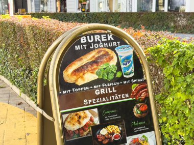 Graz, Avusturya - 11 Ekim 2025: Burek mit Joghurt tabelası renkli yeşilliklerle çevrili canlı bir açık alanda ızgara spesiyaliteleri vurguluyor