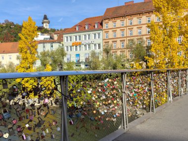 Graz, Avusturya - 11 Ekim 2025: Aşk kilitleri, renkli ağaçlar ve tarihi mimari ile çevrili, sonbaharda köprü korkuluklarını dekore eder, büyüleyici bir sahne yaratır