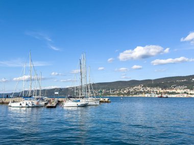 Trieste, İtalya - 14 Ekim 2025: Açık mavi gökyüzünün altında sakin bir limana demirlemiş yelkenliler, arka planda görünen tepeler, sakin bir atmosfer yaratıyor.