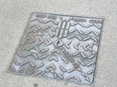 Trieste, İtalya - 14 Ekim 2025: Metal rögar kapağı detaylı desenler ve markalaşma özellikleri içeriyor, desenli beton kaldırıma yerleştirilmiş, kentsel tasarımı vurguluyor