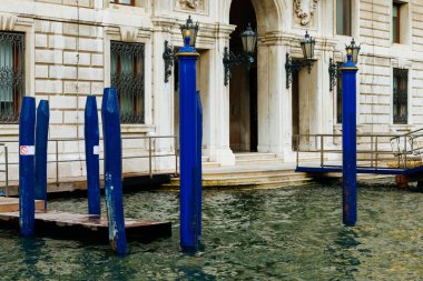 Venedik, İtalya - 23 Haziran 2025: Venedik 'teki tarihi bir binanın yanındaki sudan yükselen mavi kutuplar, kentin mimari güzelliğini ve sakin atmosferini yansıtıyor