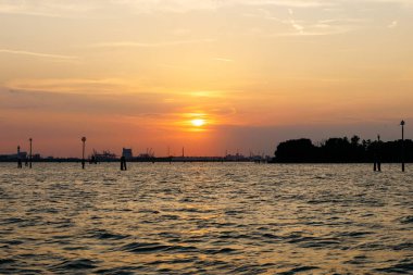 Venedik, İtalya - 23 Haziran 2025: Güzel günbatımı, ağaçların ve yapıların siluetleri ile su üzerinde ılık renkler bırakır.