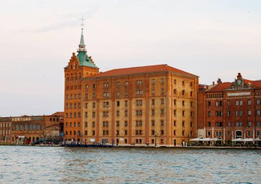 Venedik, İtalya - 23 Haziran 2025: Deniz kenarında yeşil kuleye sahip tarihi tuğla yapı, mimari ayrıntıları ve kentsel atmosferi vurguluyor