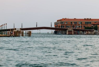 Venedik, İtalya - 23 Haziran 2025: Sahra kıyısı, sakin suları ve huzurlu bir atmosferi olan büyüleyici bir binaya açılan ahşap bir köprüye sahip.