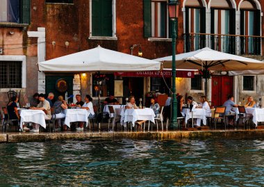 Venedik, İtalya - 23 Haziran 2025: Kanal boyunca manzaralı bir Venedik restoranında beyaz masa örtüleri ve yemeklerinin tadını çıkaran misafirler