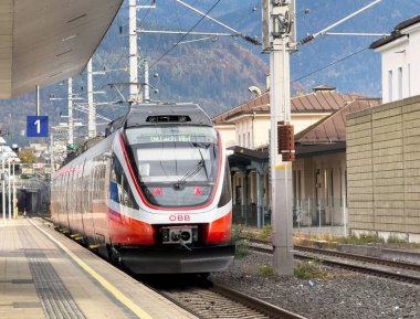 Villach, Avusturya - 23 Ekim 2025: Modern OBb treni çarpıcı dağ manzarası ve altyapısıyla çevrili istasyona geliyor.