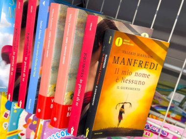 Tarvisio, İtalya - 17 Ekim 2025: Raftaki canlı İtalyan kitap koleksiyonu Valerio Massimo Manfredi 'nin başlıklarını ve rahat bir kitapçıdaki renkli kapakları vurguluyor