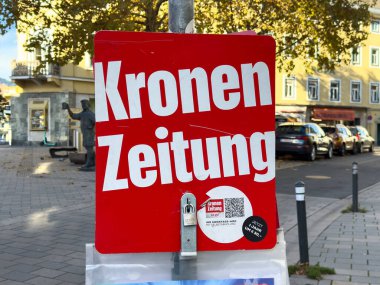 Villach, Avusturya - 29 Ekim 2025: Parlak kırmızı işaret Kronen Zeitung reklamını park etmiş arabalar ve ağaçlarla dolu canlı kentsel bir ortamda gösteriyor