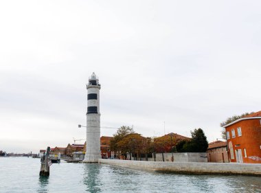 Murano, İtalya - 21 Kasım 2024: Murano adasındaki deniz feneri, sakin sular ve sonbahar yapraklarıyla çevrili Venedik mimarisini yansıtıyor ve ziyaretçileri bölgeyi keşfetmeye çağırıyor