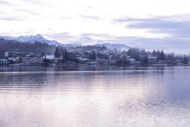 Velden, Avusturya - 20 Aralık 2024: Schlosshotel am Woerthersee kar kaplı dağlar ve sakin göl ile bir kış manzarası sergiliyor.