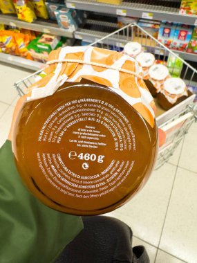 Villach, Avusturya - 25 Kasım 2025: Elindeki meyve reçeli kavanozu markette turuncu kapaklı, ürün bilgilerini ve canlı ambalajı vurguluyor