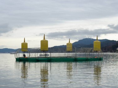 Velden, Avusturya - Aralık 06, 2025: Yüzen balık çiftliği, sakin su üzerinde sarı şamandıralar ve yeşil ağlar barındırıyor.