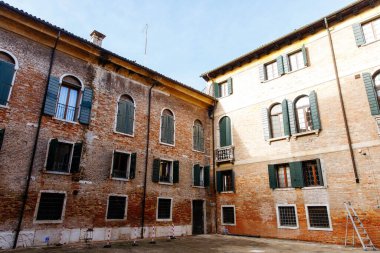 Venedik, İtalya - 05 Aralık 2025: Yeşil panjurlu tarihi tuğla yapılar, mimari güzelliği ve davetkar atmosferi ön plana çıkaran parlak bir avlu oluşturuyor