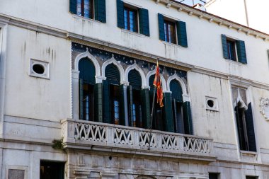 Venedik, İtalya - 05 Aralık 2025: Karmaşık pencereleri, yeşil kepenkleri ve renkli bir bayrağı olan tarihi Venedik binası cephesi, mimari zarafet ve kültürel önemi vurguluyor