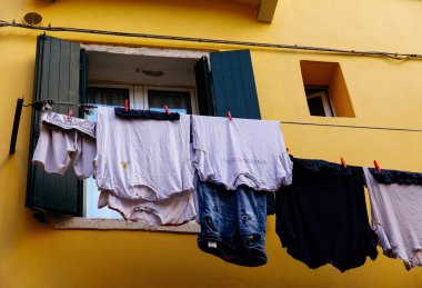 Venedik, İtalya - 05 Aralık 2025: Renkli giysiler pencerenin dışındaki çamaşır ipinde asılı, parlak sarı duvarlı bir ortam yaratıyor.