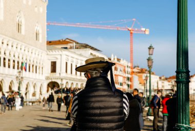 Venedik, İtalya - 05 Aralık 2025: Şapkalı bir adam Venedik 'teki Doge Sarayı' nın önünde duruyor, çevresindeki hareketli kalabalığı ve inşaat faaliyetlerini izliyor.