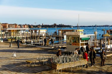 Venedik, İtalya - 05 Aralık 2025: Rıhtım sahnesinde insanlar, botlar ve şehir binaları, canlı atmosfer ve kentsel cazibe yakalayan su taksi durağı yer alıyor