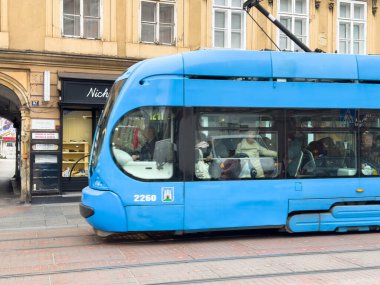 Zagreb, Hırvatistan - 14 Aralık 2025: Butik dükkanı yakınlarındaki şehir caddesi boyunca mavi tramvay seferi, hareketli bir ortamda kentsel yaşam ve toplu taşımacılığı vurguluyor