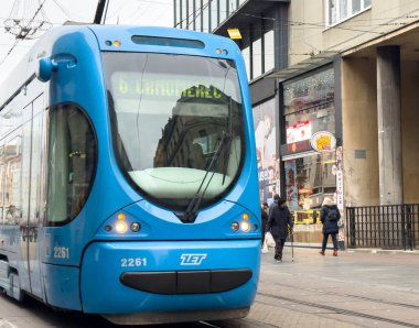 Zagreb, Hırvatistan - 14 Aralık 2025: Arka planda yayalar ve dükkanlarla toplu taşımayı ön plana çıkaran mavi tramvay şehir sokaklarında ilerliyor