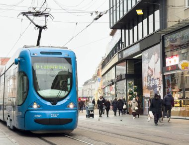 Zagreb, Hırvatistan - 14 Aralık 2025: Yayalar ve dükkanlarla dolu kentsel cadde boyunca hareket eden mavi tramvay, canlı şehir atmosferini ve toplu taşımacılığı vurguluyor