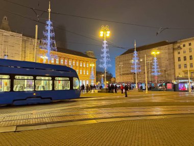 Zagreb, Hırvatistan - 14 Aralık 2025: Mavi tramvay ışıldayan ağaçlarla süslenmiş canlı bir meydanda seyahat ederek, kış kutlamalarının neşeli ruhunu gözler önüne serdi