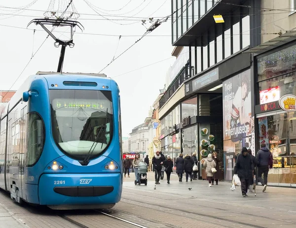 Zagreb, Hırvatistan - 14 Aralık 2025: Yayalar ve dükkanlarla dolu kentsel cadde boyunca hareket eden mavi tramvay, canlı şehir atmosferini ve toplu taşımacılığı vurguluyor